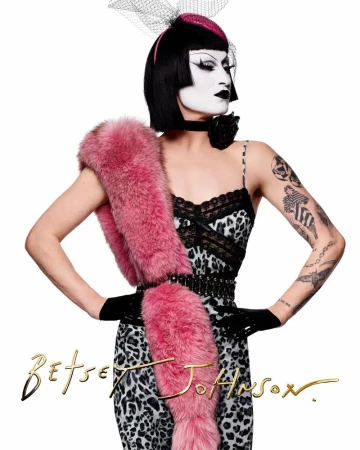 Person in striking makeup and a leopard-print dress poses with a pink fur stole in a brightly lit studio, fashion-forward and theatrical.