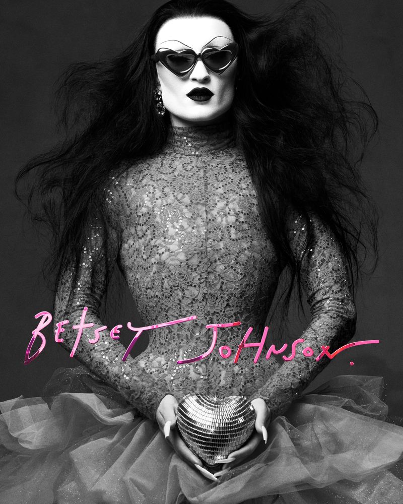 Person in heart-shaped sunglasses holding a heart disco ball, wearing a sequined turtleneck and standing against a grey backdrop.