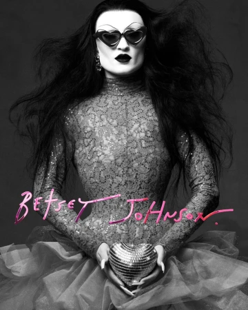Person in heart-shaped sunglasses holding a heart disco ball, wearing a sequined turtleneck and standing against a grey backdrop.