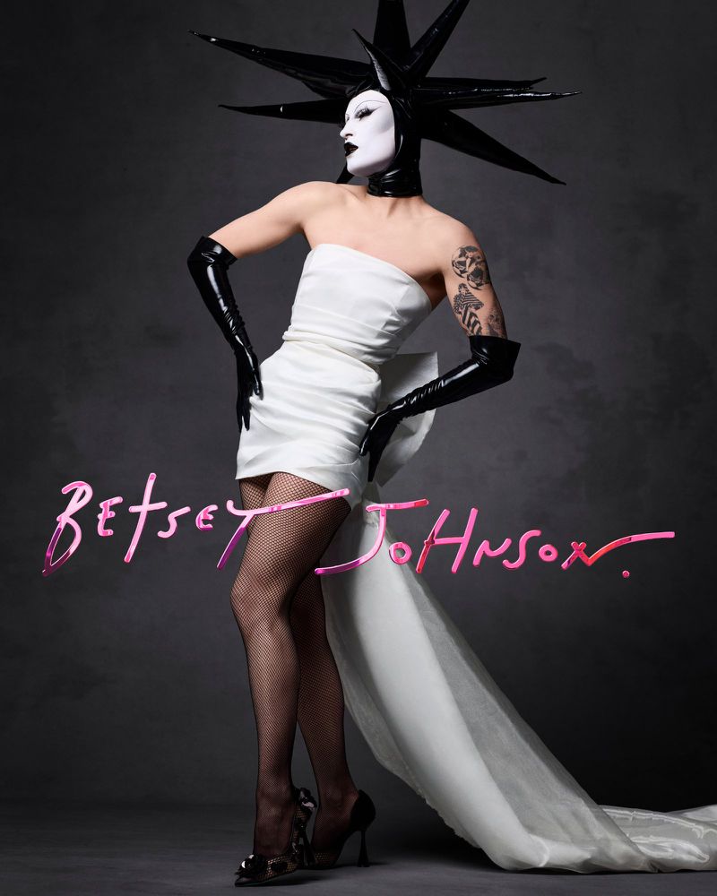 Drag queen in white dress with black headdress and gloves posing in studio with pink designer logo against gray backdrop.