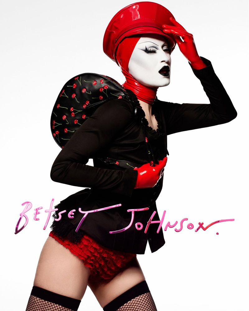 A person in theatrical makeup poses against a white backdrop, wearing a red hat, red ruffled shorts, and a black top.
