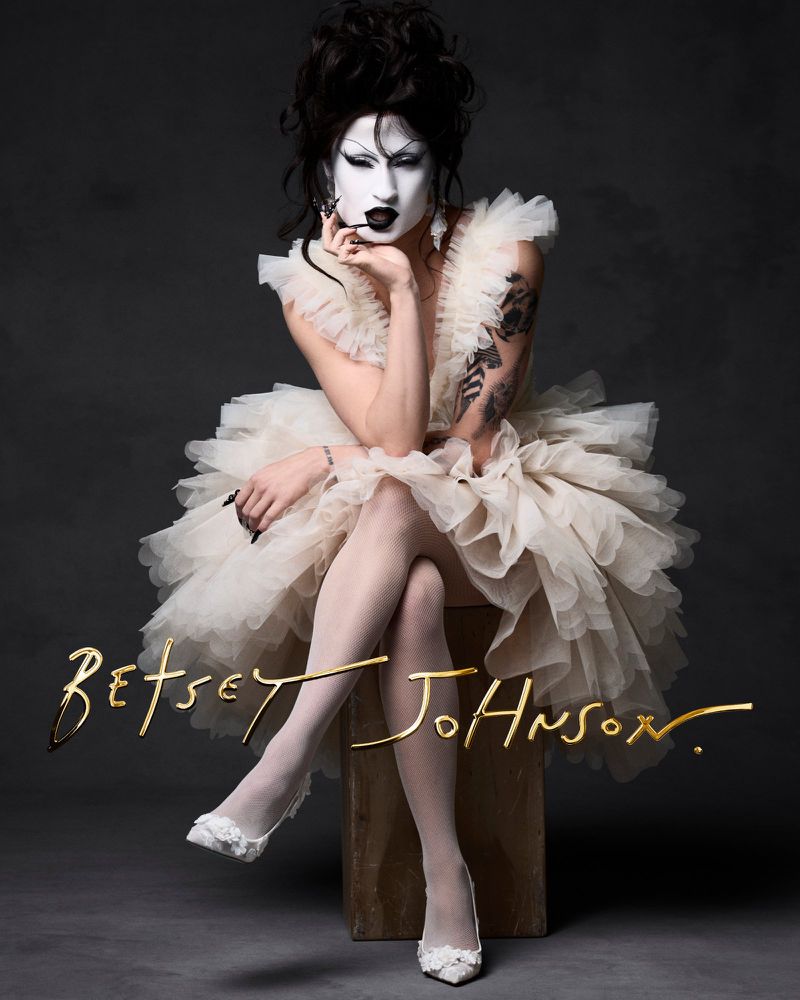 Female drag queen in a white tutu, tights, and heels poses on a wood block in a studio with a dark backdrop wearing face makeup.