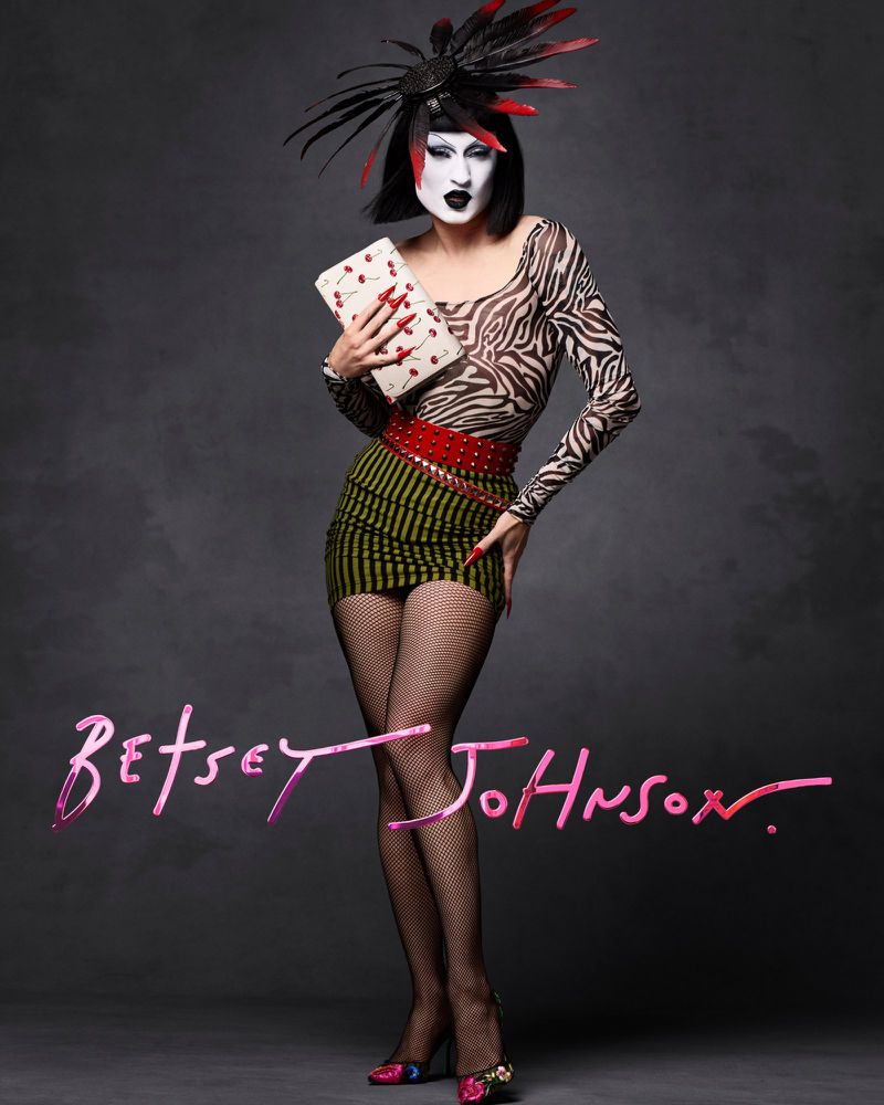 Drag queen in a zebra-print top, striped skirt, fishnet stockings, and feathered headdress, holding a cherry-patterned clutch in studio setting.