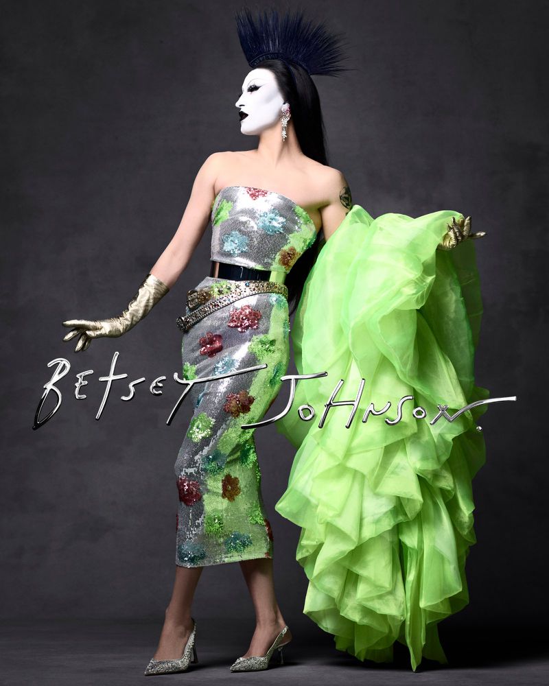 Drag queen poses in a sequined floral dress with structured shoulders, statement gloves, and an avant-garde headpiece against a grey backdrop.