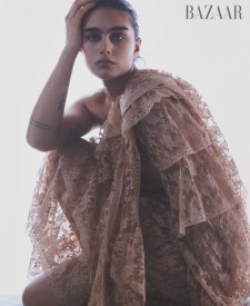 An adult female model poses in a low angle portrait in a lace gown with off-shoulder ruffles in a studio. Bazaar logo visible.