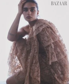An adult female model poses in a low angle portrait in a lace gown with off-shoulder ruffles in a studio. Bazaar logo visible.