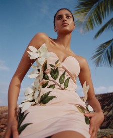 Adult woman poses wearing a light pink dress with a floral design in an outdoor setting with palm trees and a blue sky backdrop.