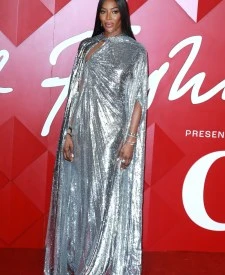 Naomi Campbell poses on a red carpet wearing a full-length metallic dress with a cape. The background is an event setting.