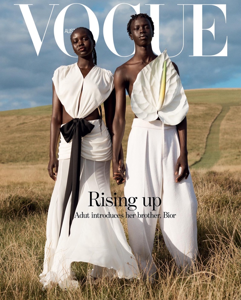 Vogue Australia cover features two dark-skinned adults in white clothing holding hands on a grassy hillside with cloudy sky backdrop.