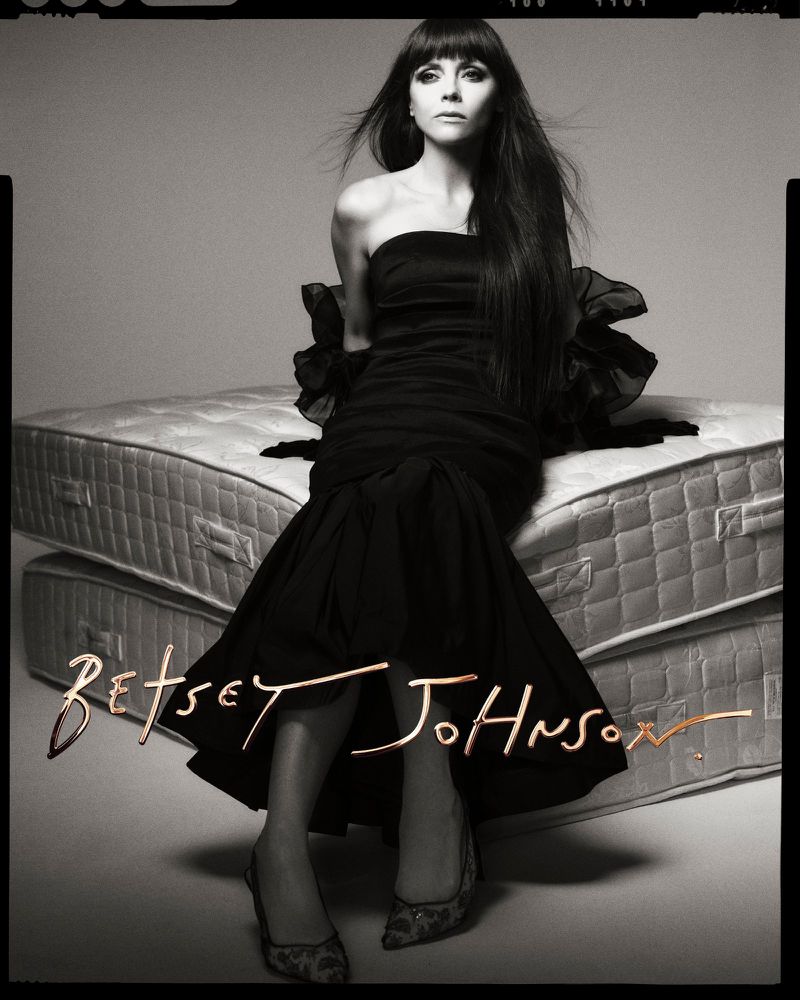A woman with long dark hair poses in a black strapless dress and heels, seated on stacked mattresses in a studio shot.
