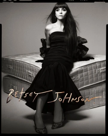 A woman with long dark hair poses in a black strapless dress and heels, seated on stacked mattresses in a studio shot.