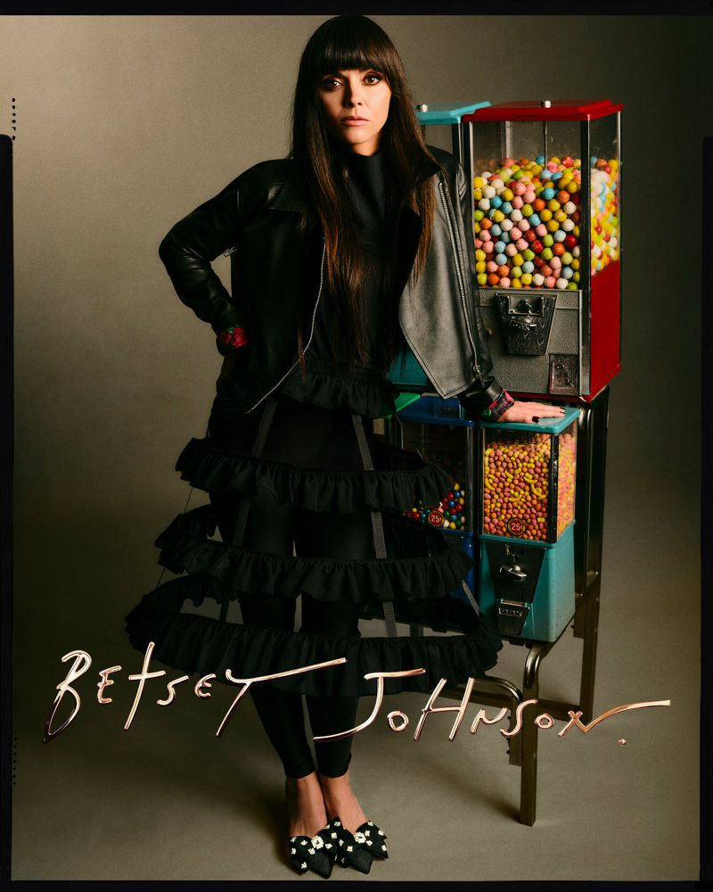 Adult woman in black outfit poses next to a candy machine in a studio with brown backdrop, showcasing fashion and unique style.