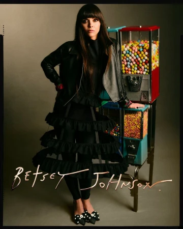 Adult woman in black outfit poses next to a candy machine in a studio with brown backdrop, showcasing fashion and unique style.