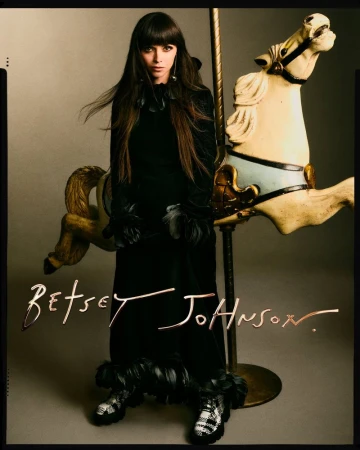 A model poses in a black dress with feather accents next to a white carousel horse in a studio setting, showcasing fashion and style.