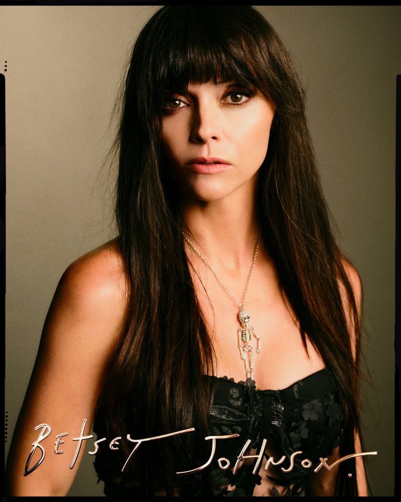 Portrait of adult woman with long dark hair, wearing black floral top and skeleton pendant, against a neutral-tone studio backdrop.
