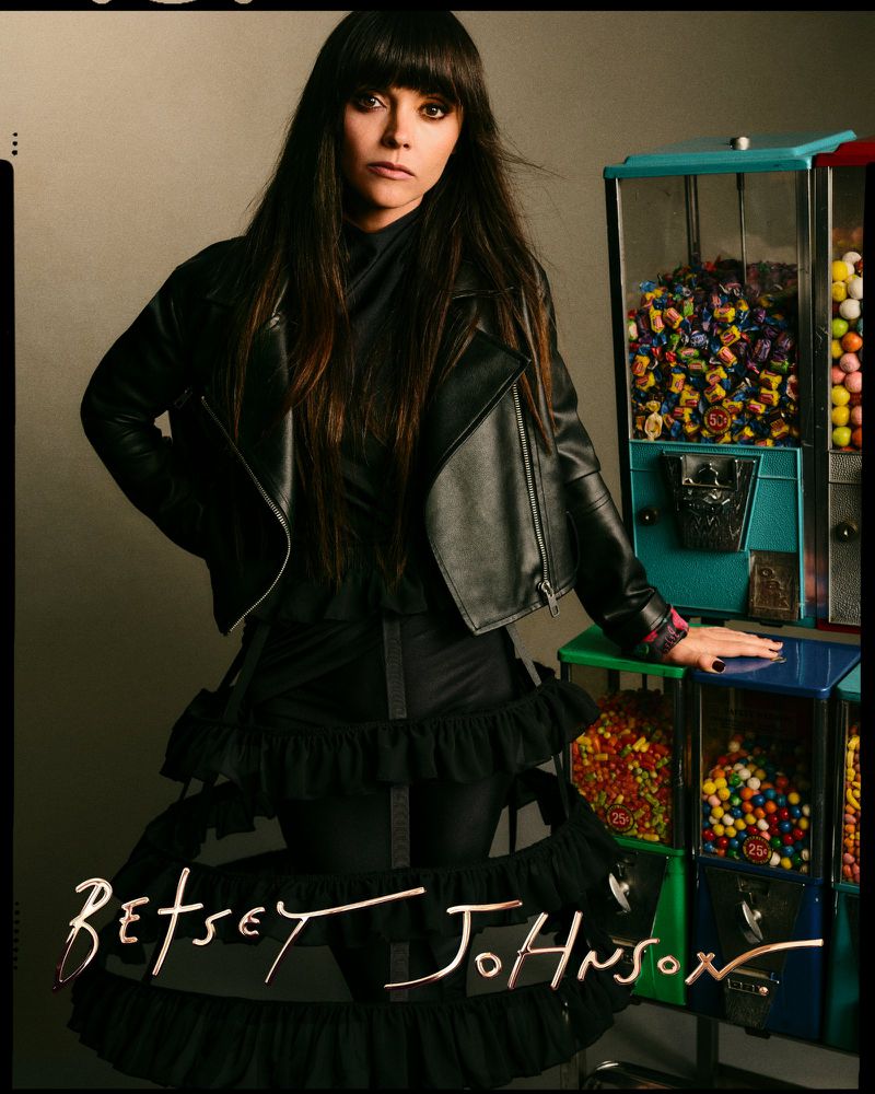 Brunette woman in a black leather jacket poses next to vintage-style candy machines, highlighted by 'Betsey Johnson' script at the bottom.