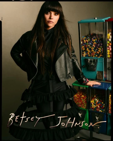 Brunette woman in a black leather jacket poses next to vintage-style candy machines, highlighted by 'Betsey Johnson' script at the bottom.
