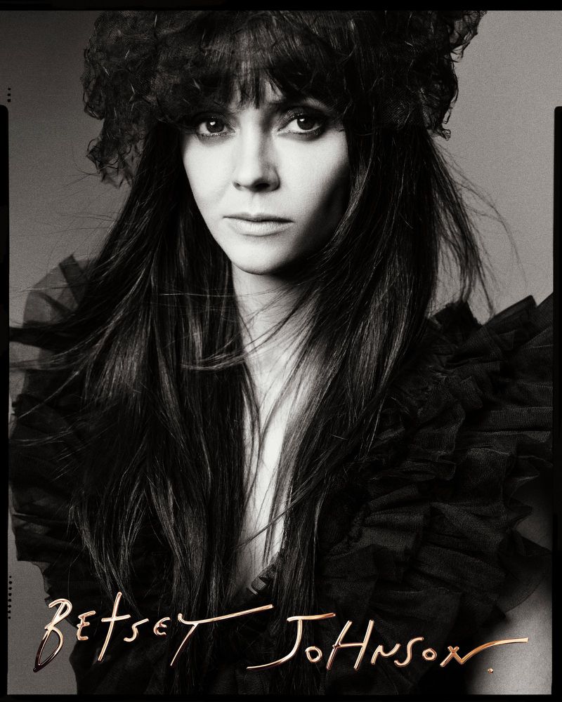 Black and white portrait of a woman with long dark hair, wearing a ruffled top and decorative headpiece, set against a neutral backdrop.