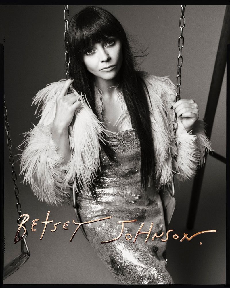 Black-and-white portrait of a female in a glittering dress and feathered coat, posing with chains, highlighting fashion and style.
