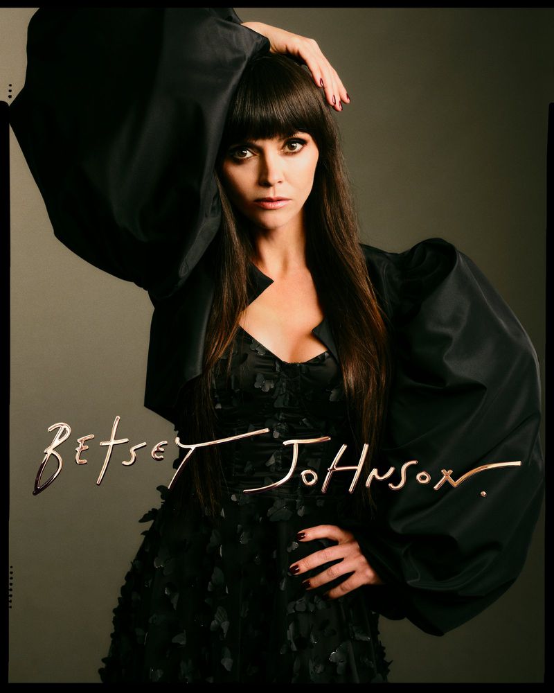 Portrait of adult woman with long brown hair and bangs wearing a black dress and jacket, posing against a grey studio backdrop.