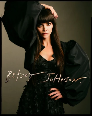 Portrait of adult woman with long brown hair and bangs wearing a black dress and jacket, posing against a grey studio backdrop.