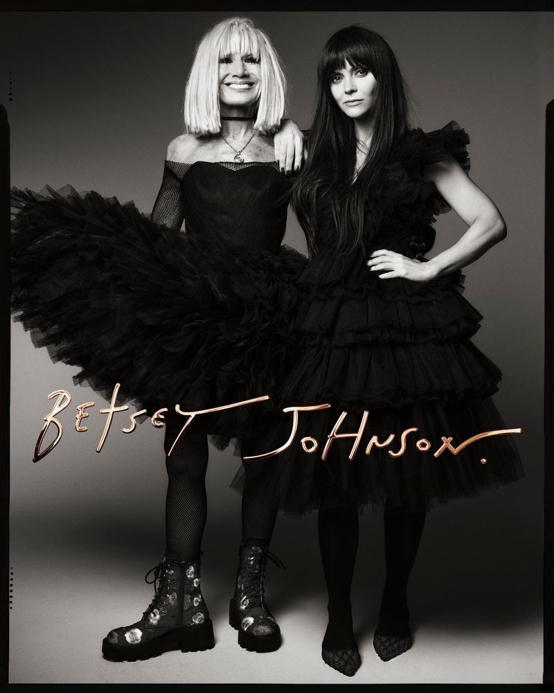 Two women in black dresses pose together in a studio shot. The woman on the left wears a large tulle skirt and platform boots.