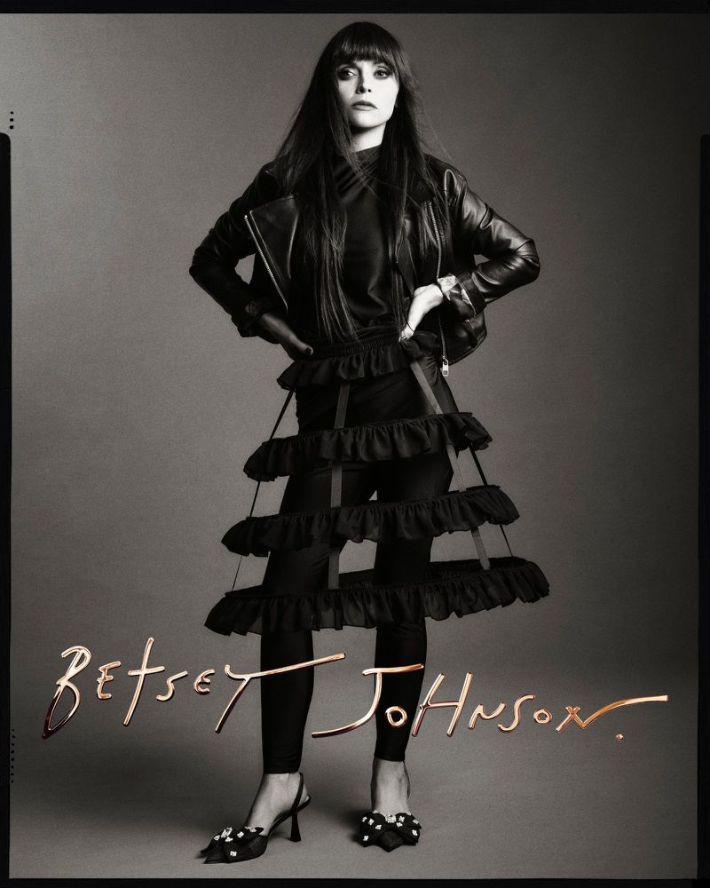 Monochrome portrait of a woman wearing a black leather jacket, tiered ruffle skirt, and heels, posing against a grey background.