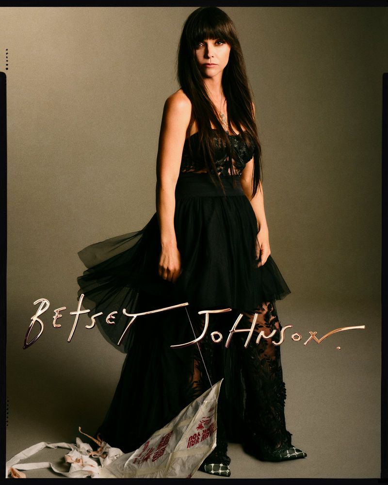 Fashion portrait of a woman in a black dress with floral details, posing in a studio with a tan backdrop and signature text overlay.