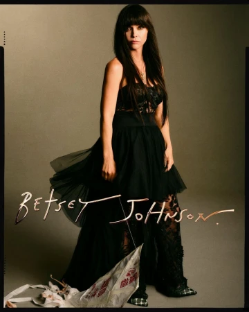 Fashion portrait of a woman in a black dress with floral details, posing in a studio with a tan backdrop and signature text overlay.