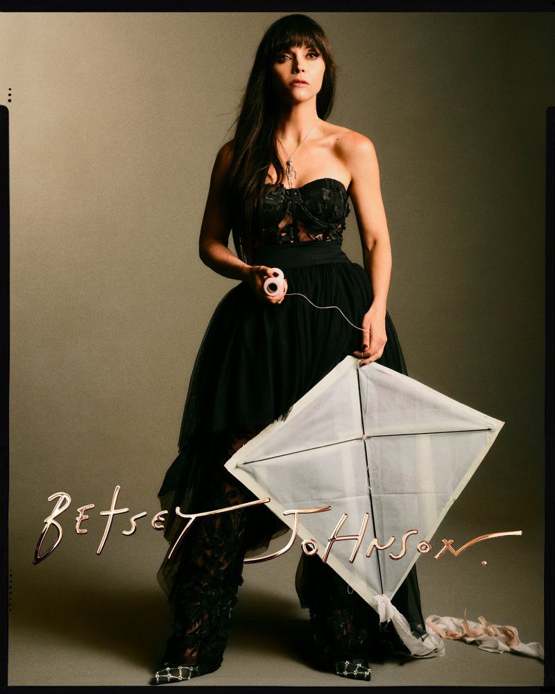 Adult woman in an elegant black dress holds a white kite in a studio setting, with the fashion brand Betsey Johnson visible.