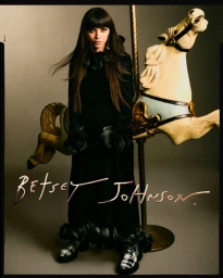 Christina Ricci standing beside a vintage carousel horse in a black feathered gown and plaid platform boots for Betsey Johnson.