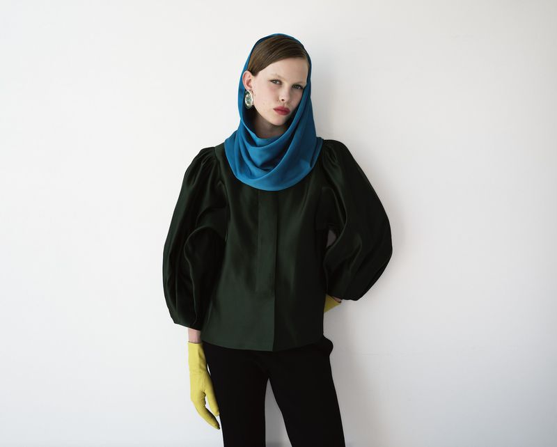 Elegant studio shot of a stylish woman wearing a blue headscarf, dark green blouse with voluminous sleeves, black pants, and yellow gloves.