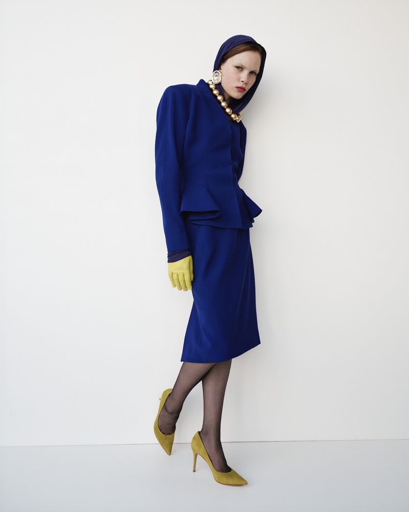 A woman in a blue skirt suit, pearl necklace, and yellow gloves and heels poses against a white background in a studio setting.