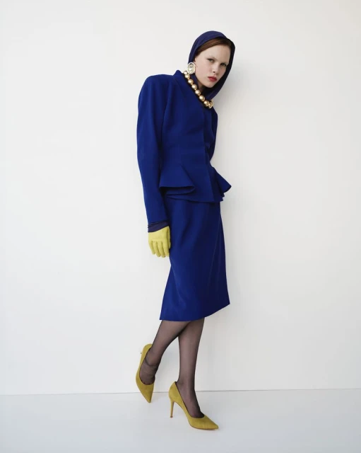 A woman in a blue skirt suit, pearl necklace, and yellow gloves and heels poses against a white background in a studio setting.