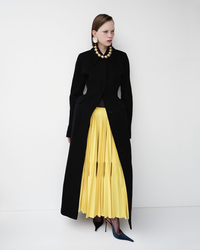 Full-length fashion shot of a model in a black coat, yellow pleated skirt, gold necklace, and blue heels against a white backdrop.