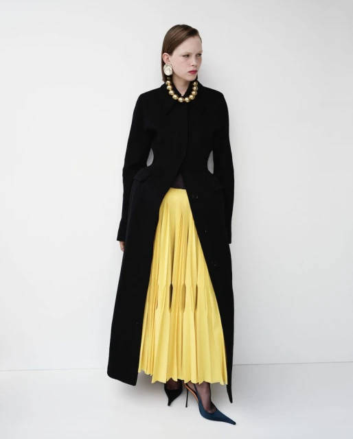 Full-length fashion shot of a model in a black coat, yellow pleated skirt, gold necklace, and blue heels against a white backdrop.