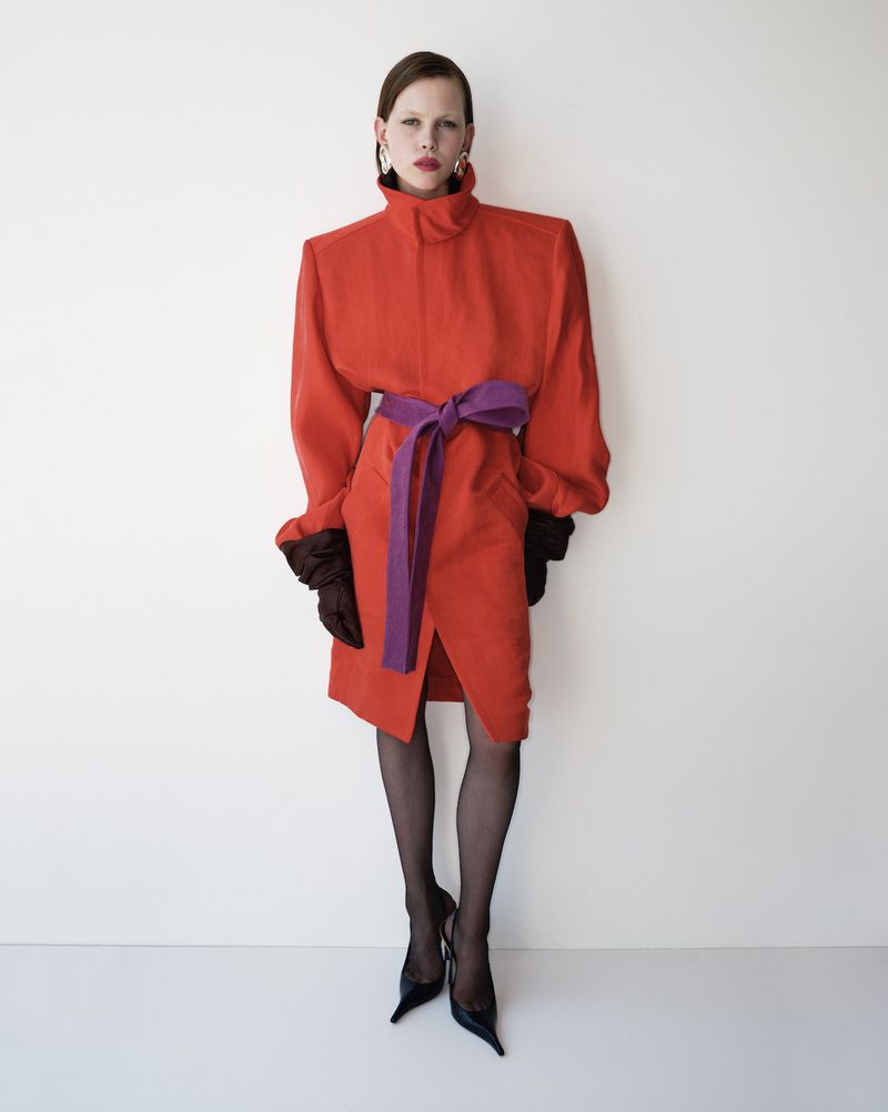 A fashionably dressed woman in a red dress cinched with a purple belt, accessorized with earrings, gloves, hose, and heels poses indoors.