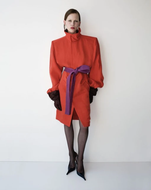 A fashionably dressed woman in a red dress cinched with a purple belt, accessorized with earrings, gloves, hose, and heels poses indoors.