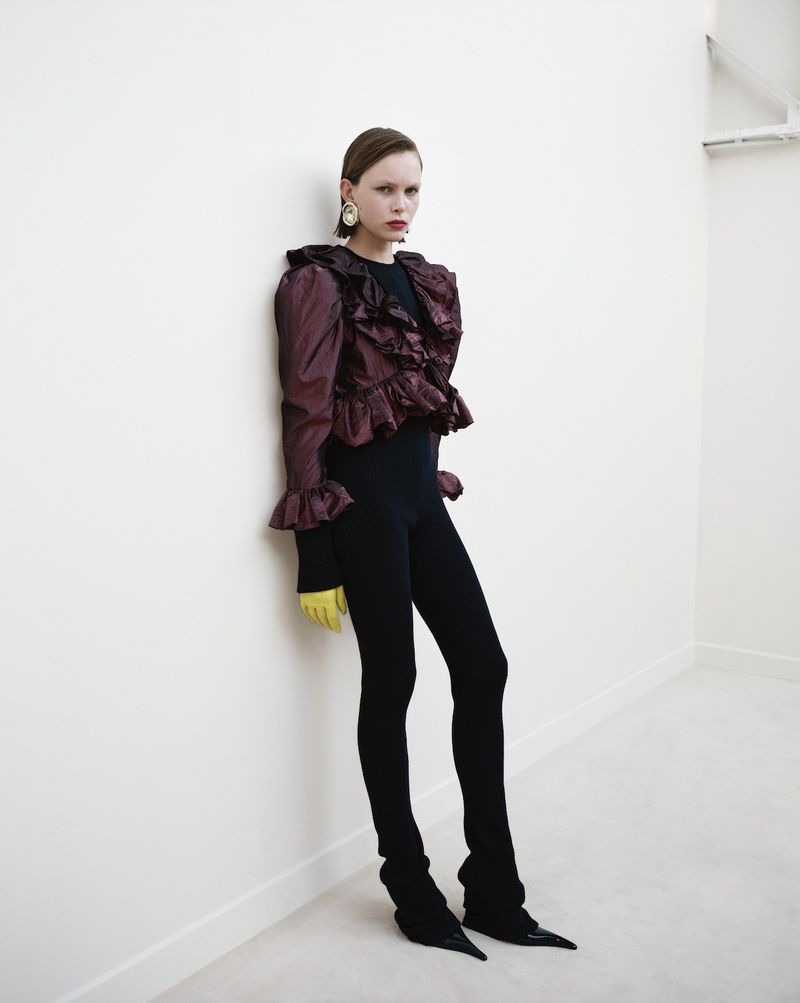 A stylish adult female model posing indoors wearing a ruffled auburn jacket, black leggings and heels with yellow gloves against a white wall.