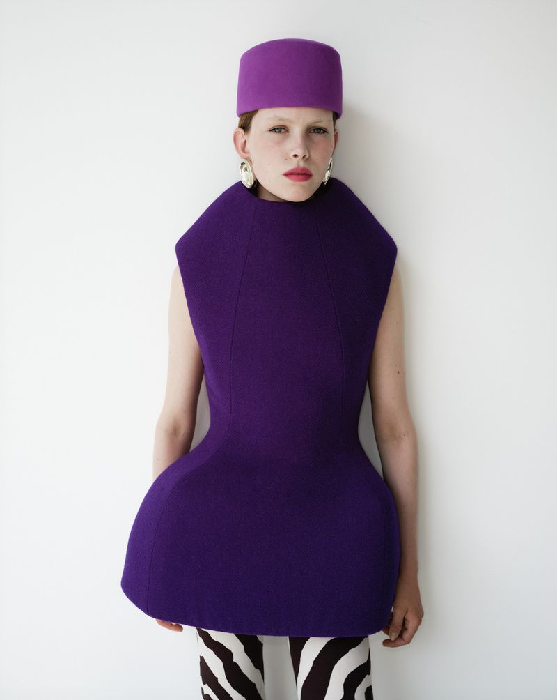 A female fashion model in a purple dress and hat stands against a white background in a studio environment.