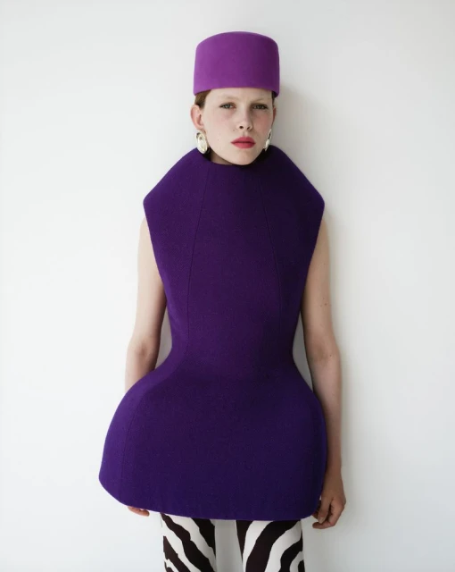 A female fashion model in a purple dress and hat stands against a white background in a studio environment.