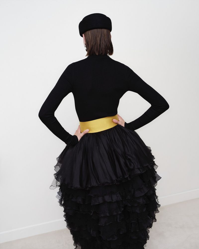 A woman poses from the back, wearing a black long-sleeved shirt, a black ruffled skirt with a gold waistband, and a black beret.