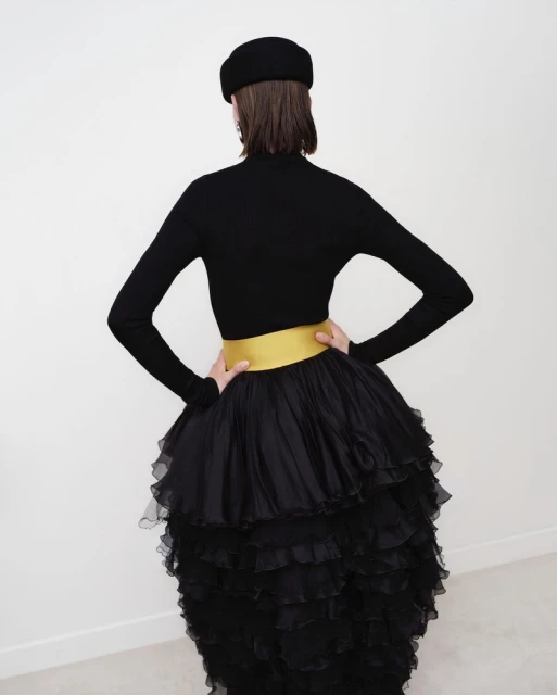 A woman poses from the back, wearing a black long-sleeved shirt, a black ruffled skirt with a gold waistband, and a black beret.