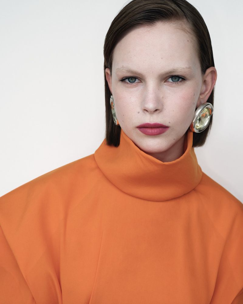 Close-up portrait of a young woman with blue eyes and short brown hair wearing an orange turtleneck and silver earrings in a studio.