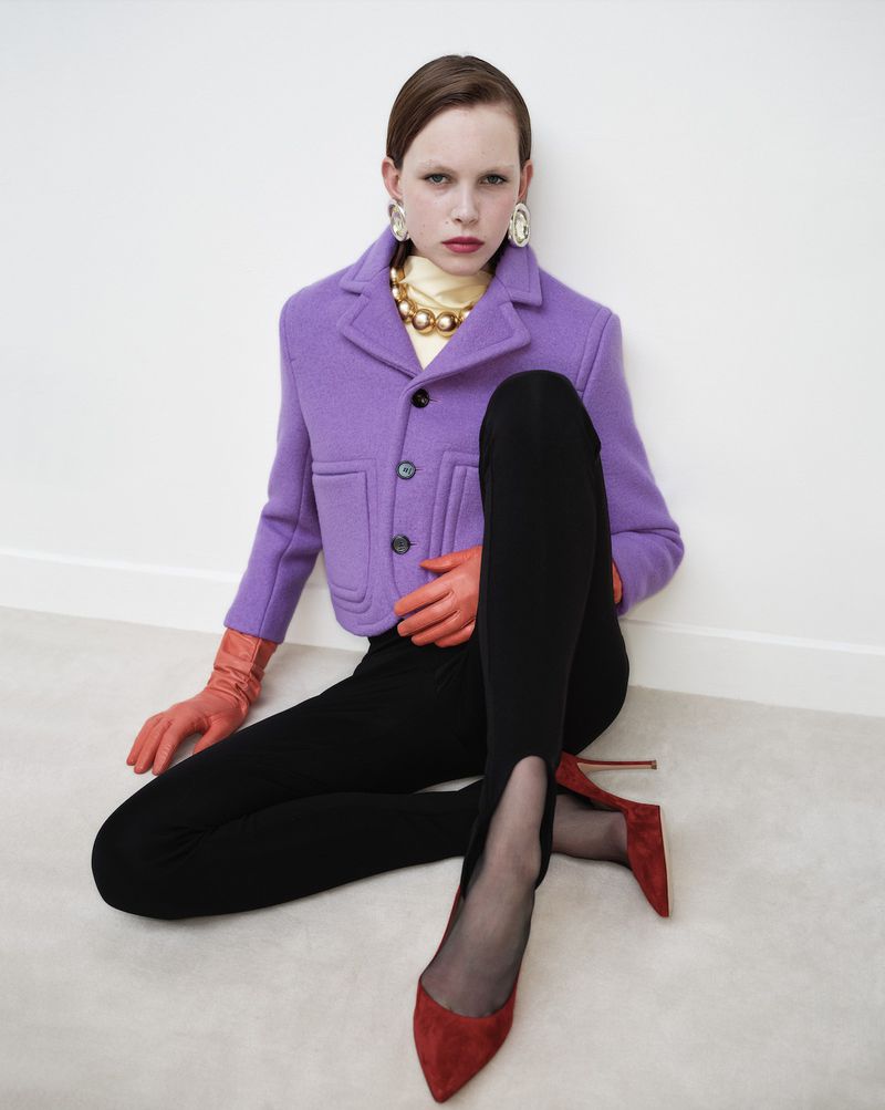 Fashionable woman with purple jacket, black leggings, and red heels poses in a minimalist white studio, showcasing stylish and modern apparel.