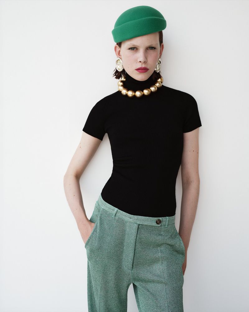 A woman in a black top and green pants stands against a white wall, accessorized with a green hat and gold necklace and earrings.