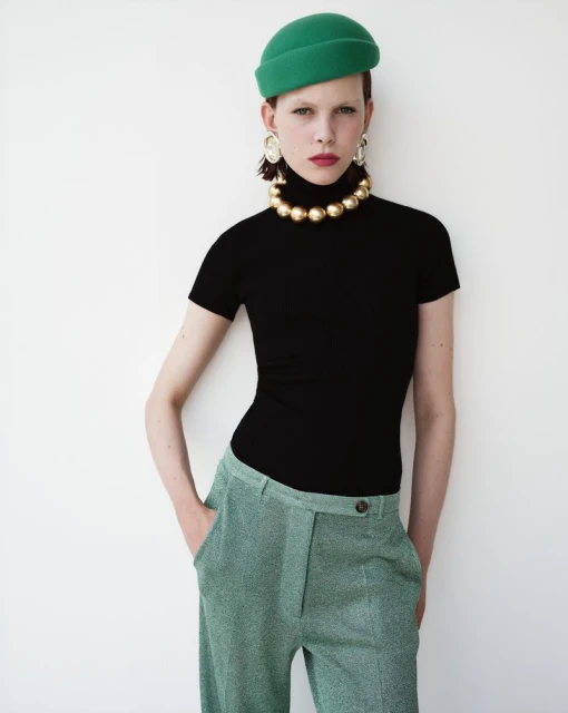 A woman in a black top and green pants stands against a white wall, accessorized with a green hat and gold necklace and earrings.
