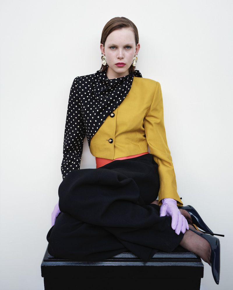 Fashion model in a polka dot and mustard yellow blazer, sitting on a black trunk against a plain background in a fashion editorial.