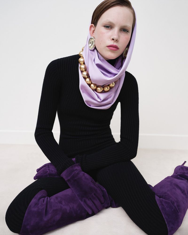 A stylish woman poses seated indoors wearing a black ribbed dress, purple boots and gloves, and a draped lilac headscarf with gold beaded necklace.