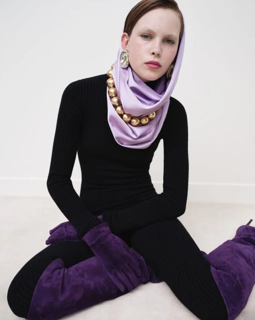 A stylish woman poses seated indoors wearing a black ribbed dress, purple boots and gloves, and a draped lilac headscarf with gold beaded necklace.